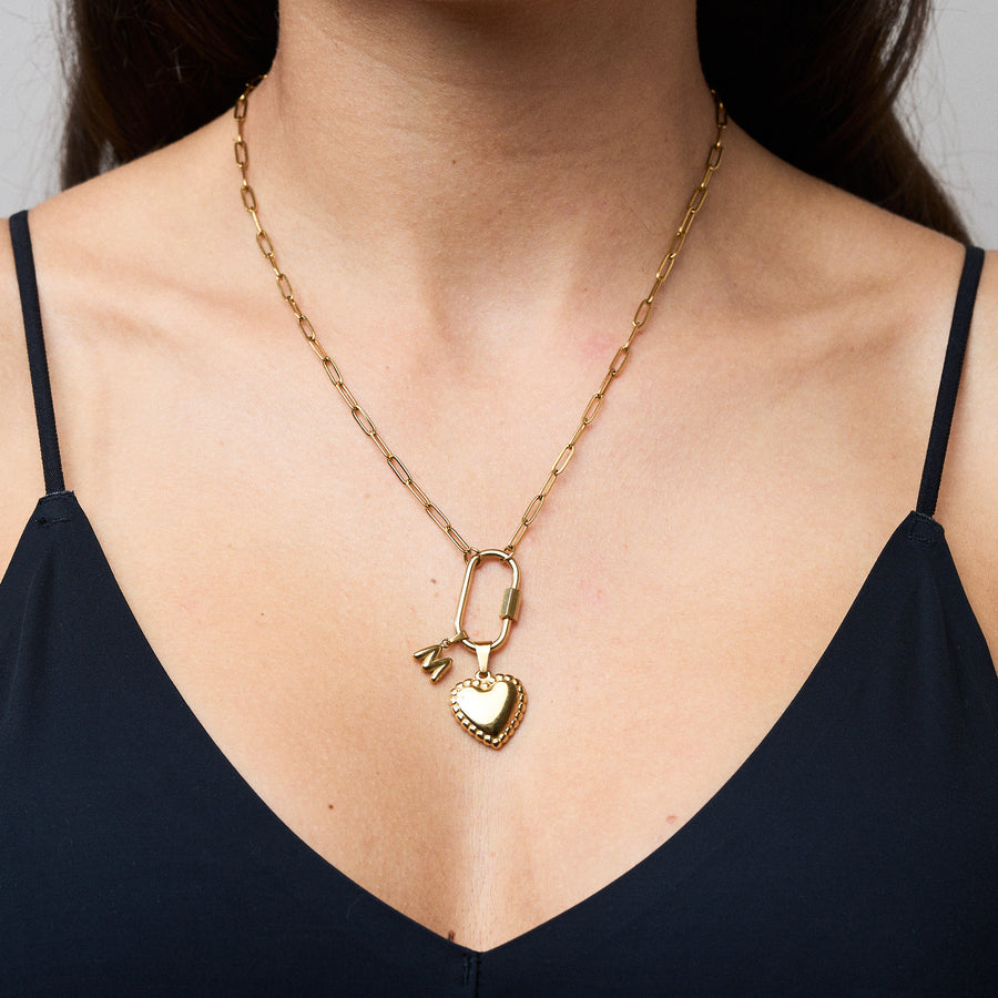 Goldene Gliederkette mit großem Karabinerverschluss, an dem ein herzförmiger Anhänger mit Perlenrand und ein kleiner Buchstabe „M“ hängen. Oben rechts steht in einem schwarzen Kasten: „Set Deal – 15 € off