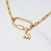 Goldene Gliederkette mit großem Karabinerverschluss, an dem ein herzförmiger Anhänger mit Perlenrand und ein kleiner Buchstabe „M“ hängen. Oben rechts steht in einem schwarzen Kasten: „Set Deal – 15 € off