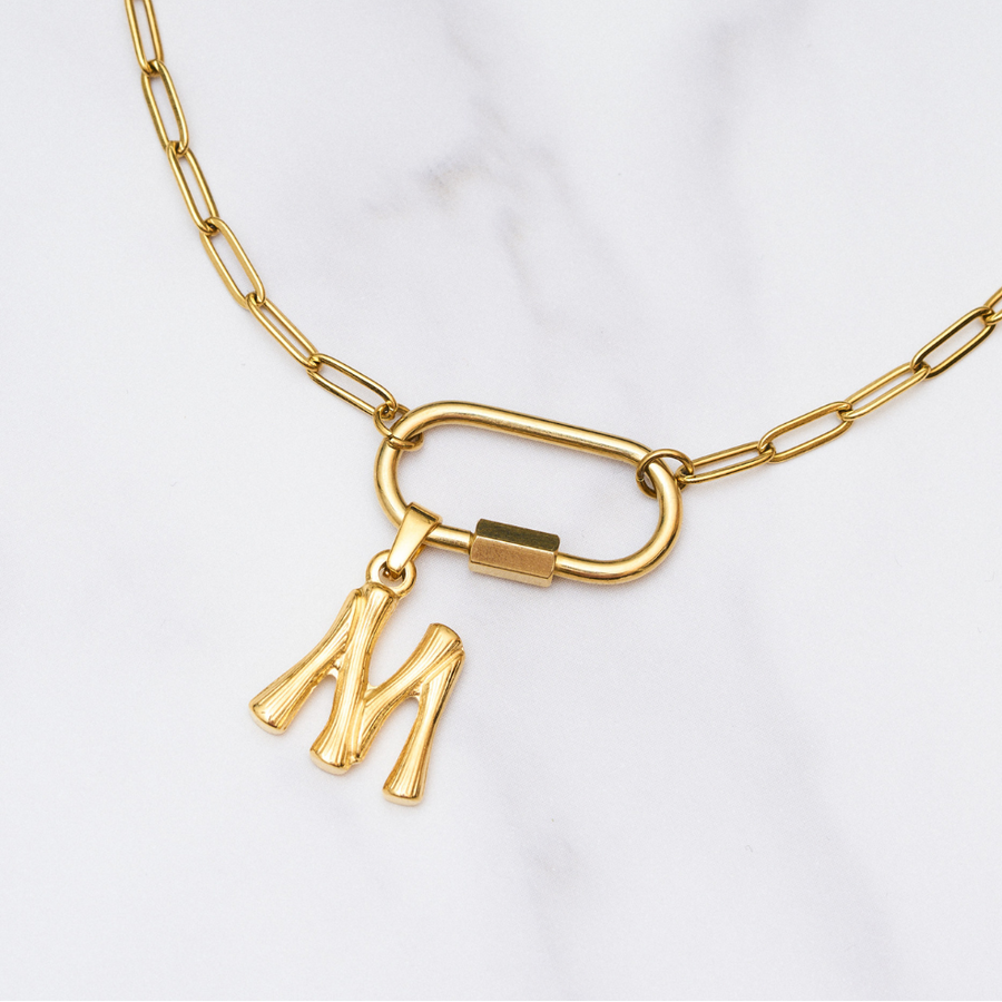 Frau trägt eine goldene Gliederkette mit großem Karabinerverschluss, an dem zwei goldene Buchstabenanhänger „B“ und „M“ hängen. Sie trägt ein geripptes, fliederfarbenes Oberteil mit quadratischem Ausschnitt. Oben rechts steht in einem schwarzen Kasten: „Set Deal – 15 € off“